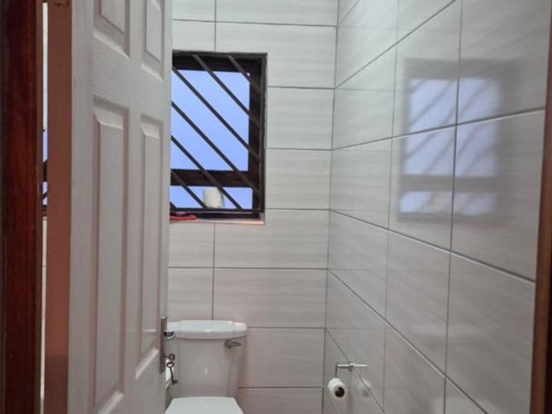 A compact, well-maintained bathroom featuring floor-to-ceiling light-toned wall tiles, tiled flooring, and a window with security bars.