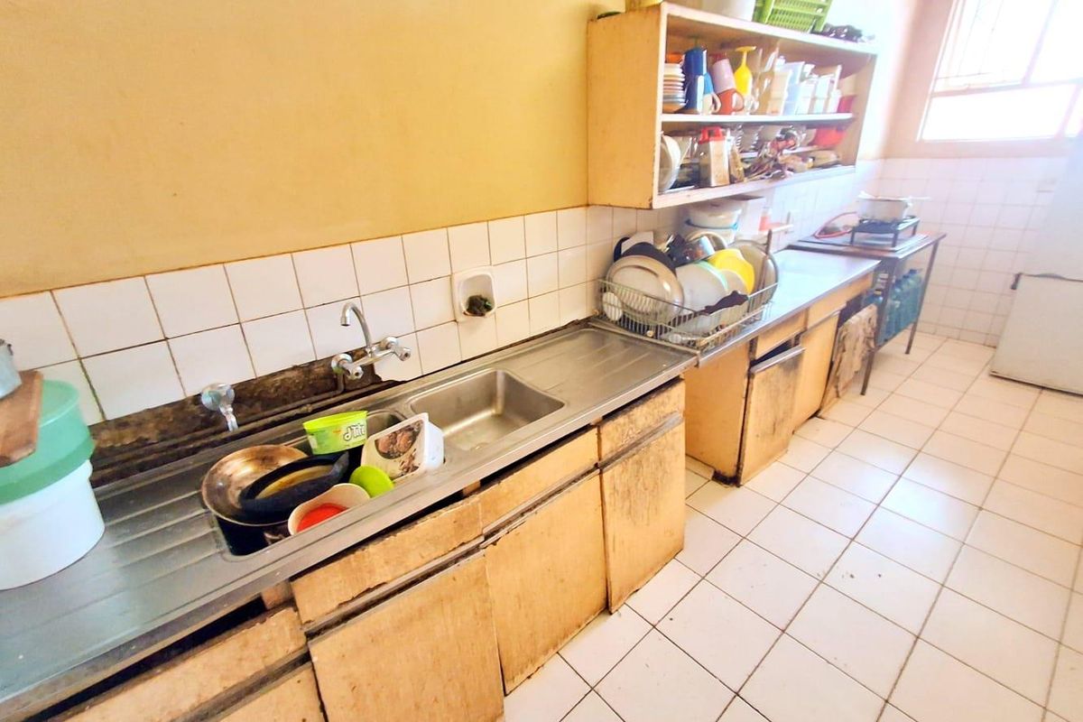 A dated kitchen featuring a stainless steel sink, tiled splashback, and worn wooden cabinetry with open shelving.