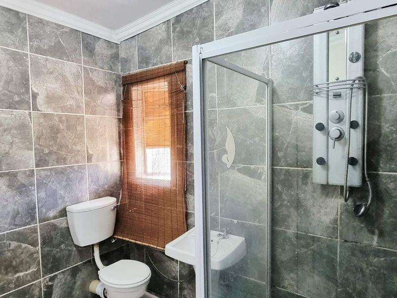 A functional bathroom featuring full-height grey stone-look wall tiles, a glass shower enclosure with a multi-jet shower panel, a toilet, and a wall-mounted basin.