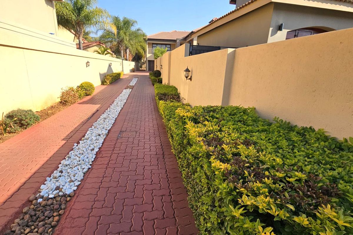 A paved side walkway featuring a central decorative stone strip, bordered by manicured hedges and high perimeter boundary walls with mounted exterior lighting.