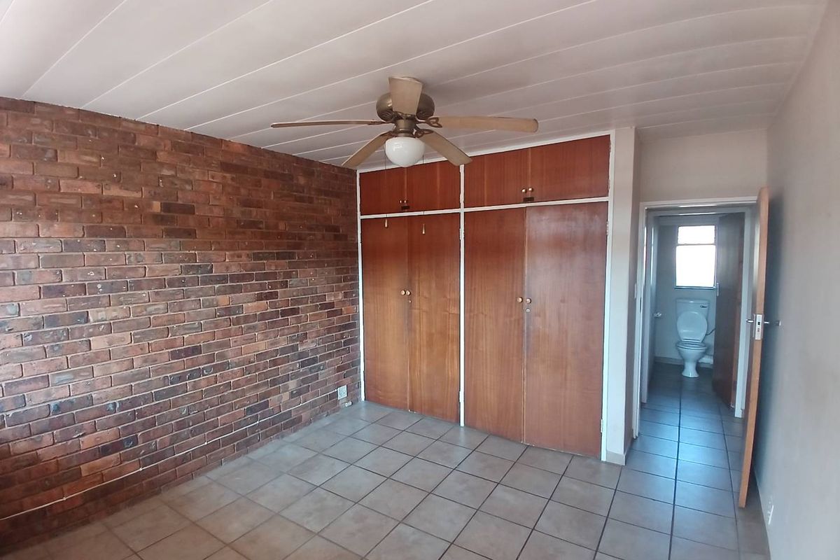 Bedroom featuring an exposed brick feature wall, built-in wooden cupboards, tiled flooring, and a ceiling fan, with an adjoining bathroom visible.