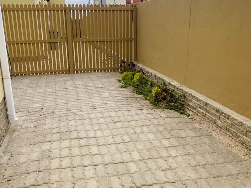 A paved rear courtyard area featuring a timber gate, boundary wall with electric fencing, and a small retaining wall with garden bed.