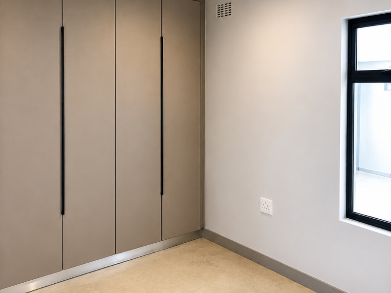 A clean, modern bedroom featuring neutral tiled flooring, built-in cupboards with sleek handles, and a large window providing natural light.
