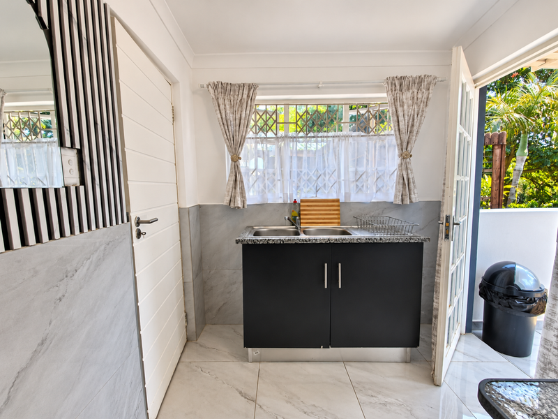 A clean and functional scullery area featuring a stainless steel double sink, granite countertop, and modern tiled finishes with direct access to an outdoor space.