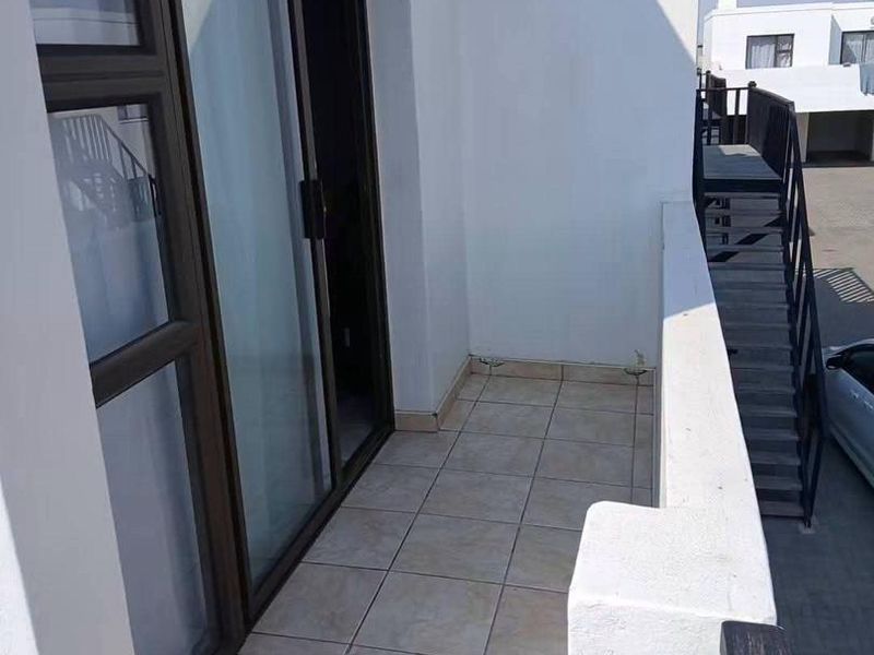 A clean, tiled balcony area featuring a sliding glass door entrance and a metal balustrade, overlooking the exterior complex grounds.