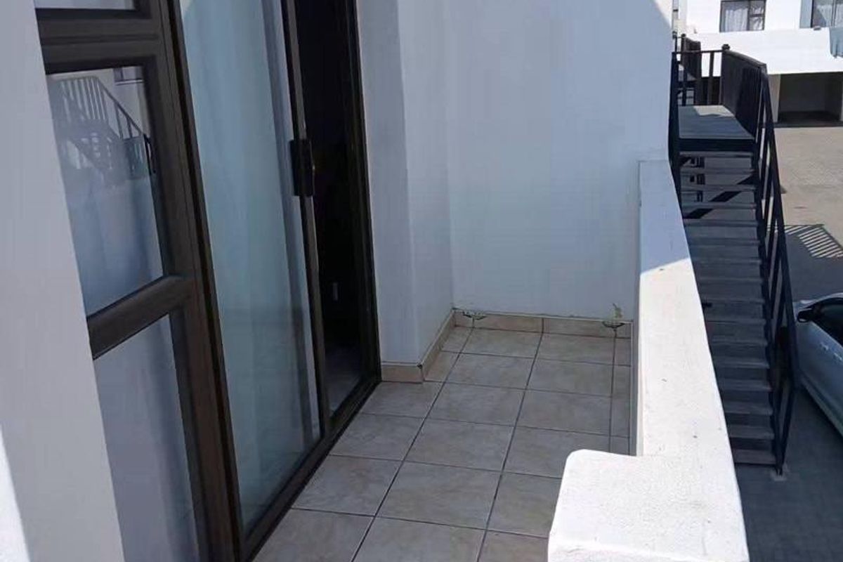 A clean, tiled balcony area featuring a sliding glass door entrance and a metal balustrade, overlooking the exterior complex grounds.