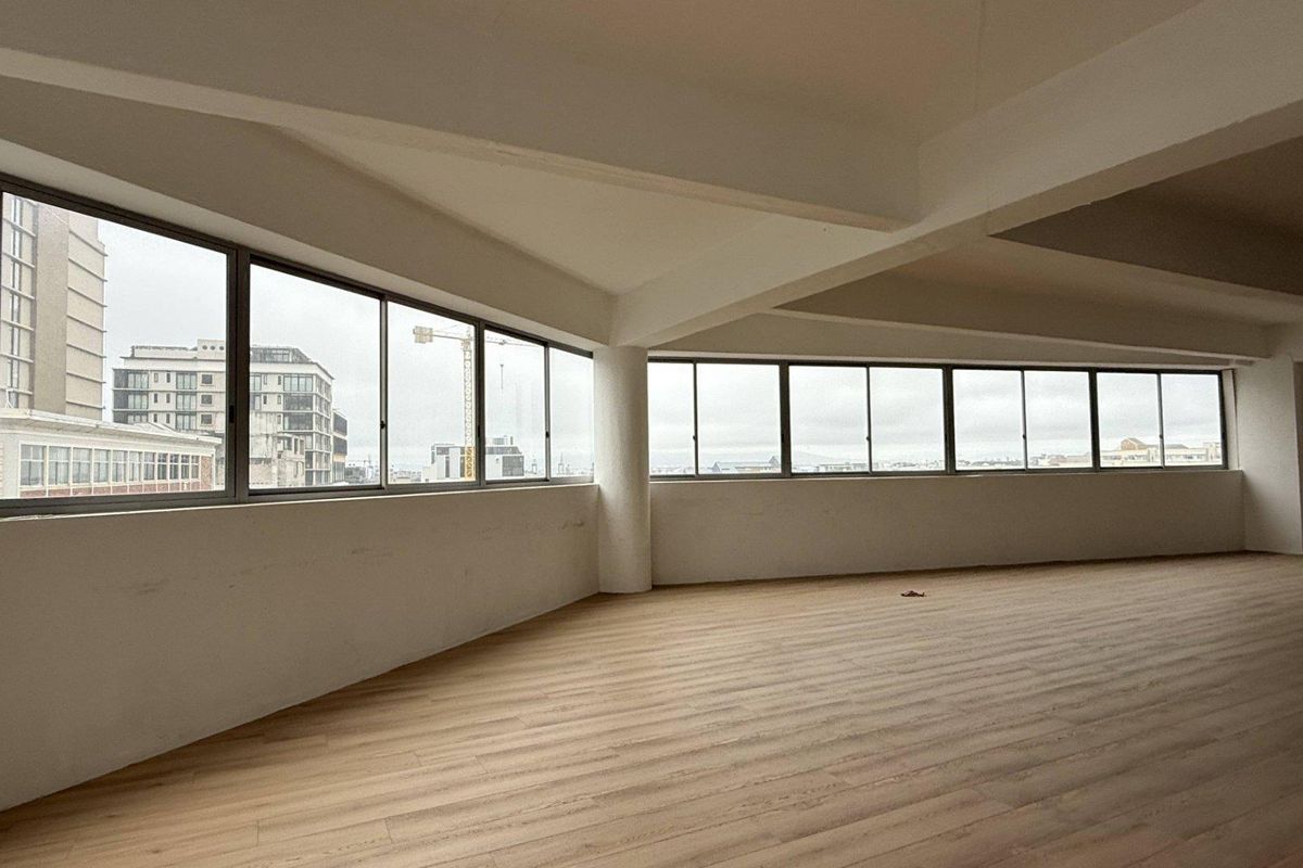 Spacious, open-plan office area featuring large windows with city views, exposed structural beams, and modern laminate flooring.