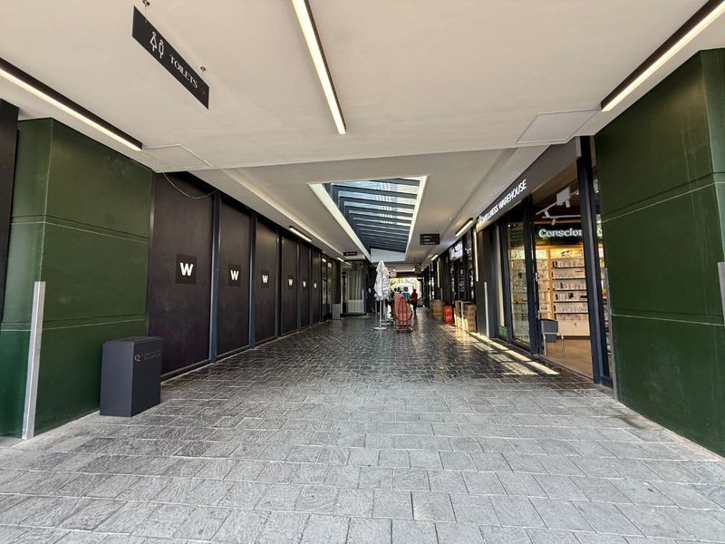 A modern, well-lit retail arcade hallway featuring stone flooring, a central skylight, and glass-fronted commercial units.