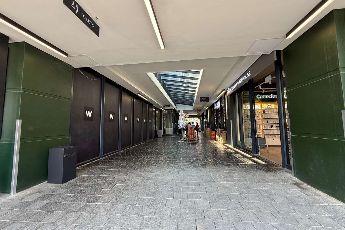 A modern, well-lit retail arcade hallway featuring stone flooring, a central skylight, and glass-fronted commercial units.