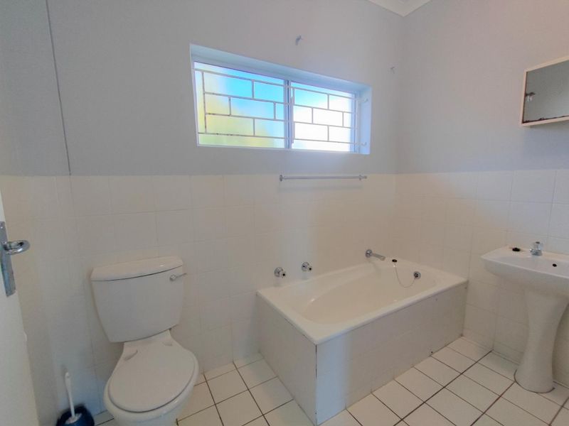 A functional bathroom featuring a built-in bathtub, pedestal basin, and toilet with neutral tiled finishes and a window with security bars.