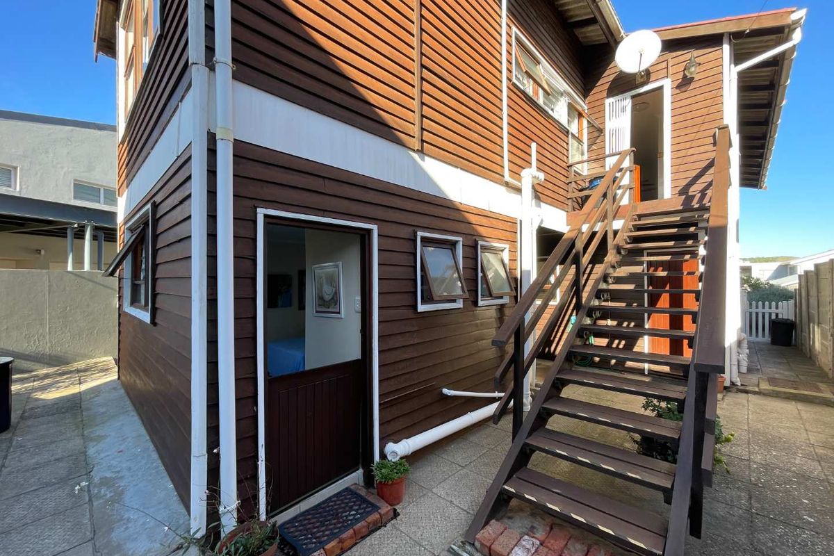 Exterior view of a two-story timber-clad house featuring an external wooden staircase leading to an upper-level entrance and a paved courtyard area.