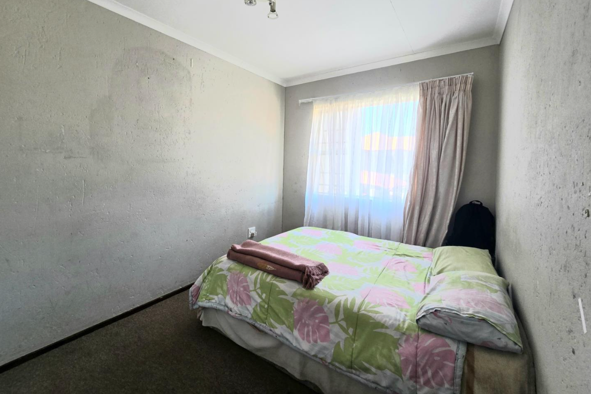 A compact bedroom featuring neutral walls, carpeted flooring, and a window with curtain fixtures, offering a functional space for rest.