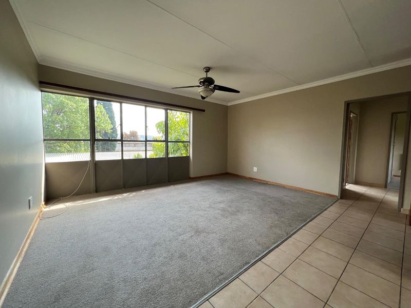 Spacious, empty living room featuring a ceiling fan, large windows for natural light, and a combination of carpet and tiled flooring.