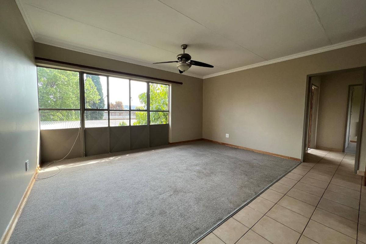 Spacious, empty living room featuring a ceiling fan, large windows for natural light, and a combination of carpet and tiled flooring.