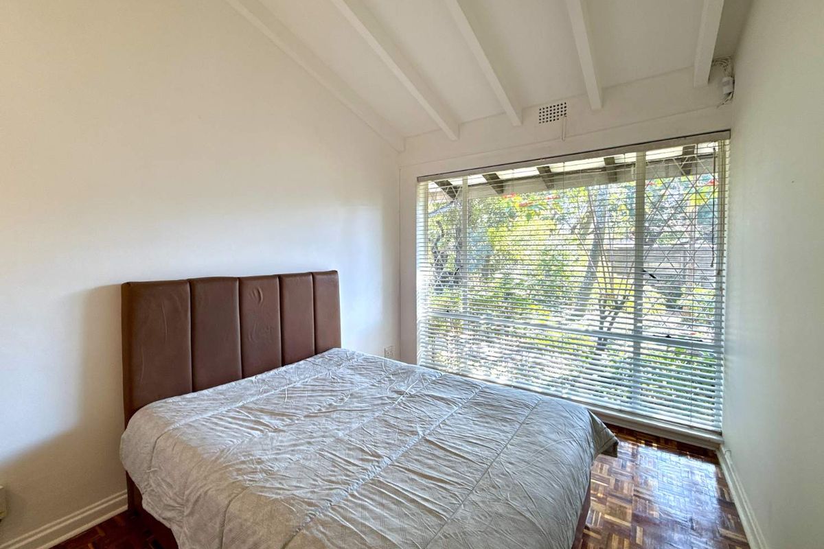 A bright bedroom featuring classic parquet flooring, exposed beam ceilings, and a large window with venetian blinds overlooking the garden.