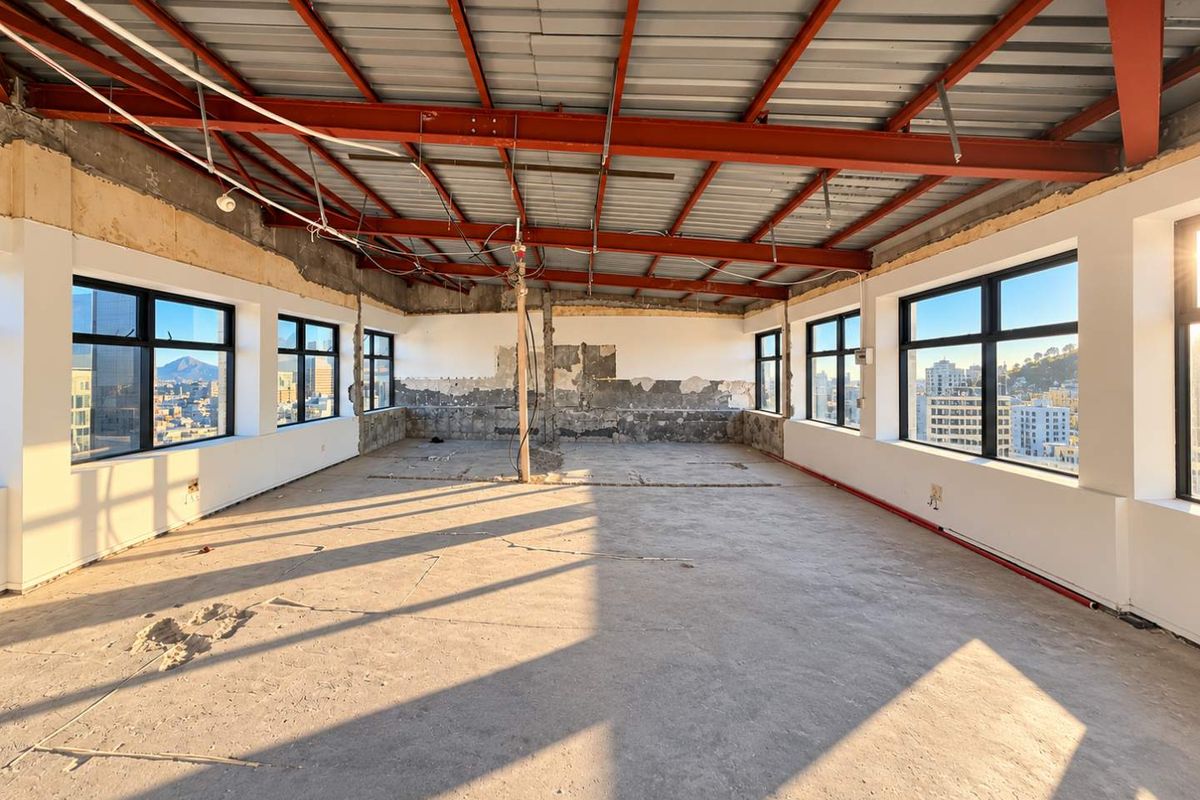 A spacious, unfinished open-plan penthouse area featuring exposed steel roof trusses, concrete floors, and large windows offering expansive city views.