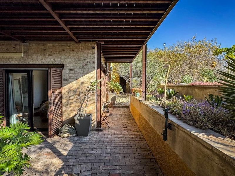 A covered outdoor patio featuring brick paving, a timber pergola, and direct access to the interior through sliding glass doors with timber shutters.