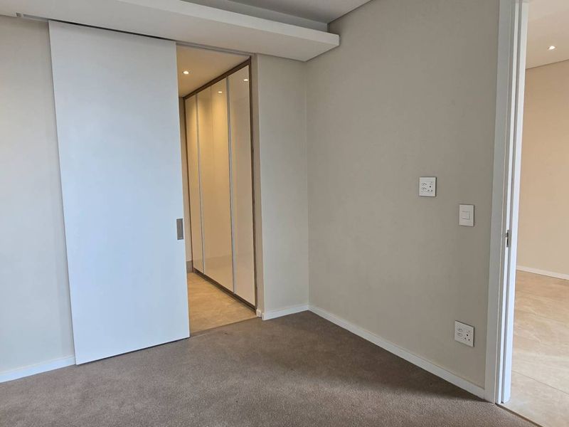 A bright, empty bedroom featuring neutral carpeting, a modern sliding door, and built-in cupboards, with an adjacent tiled hallway visible.