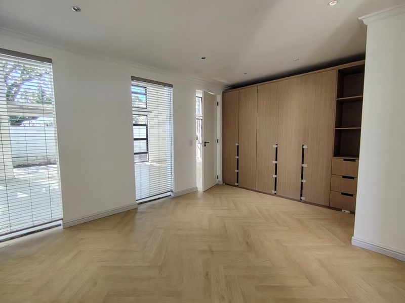 A bright, modern bedroom featuring light wood herringbone flooring, ample built-in cupboard storage, and large windows with fitted venetian blinds.