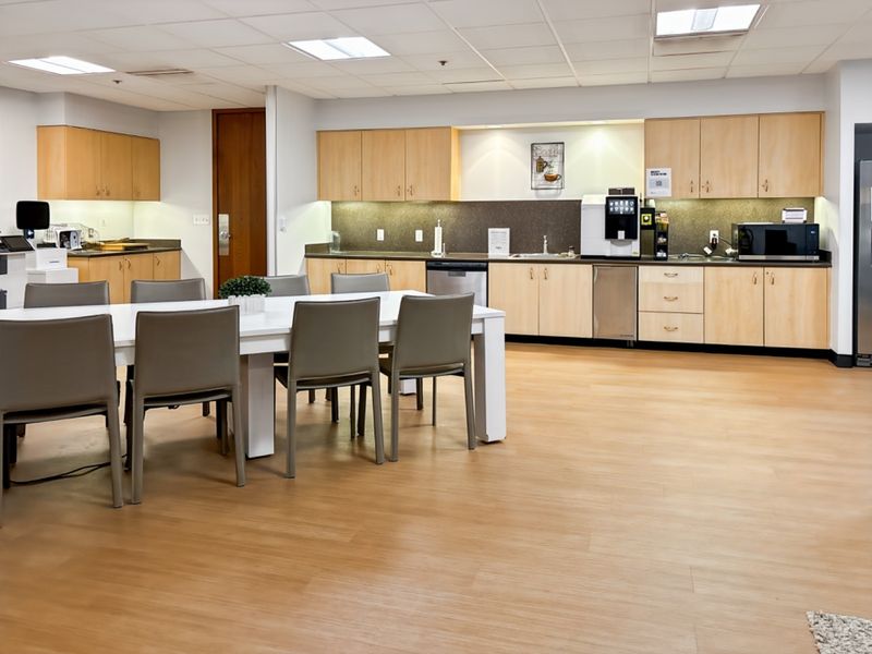 A spacious open-plan office breakroom featuring light wood cabinetry, integrated sink, modern laminate flooring, and a functional kitchenette area with recessed lighting.