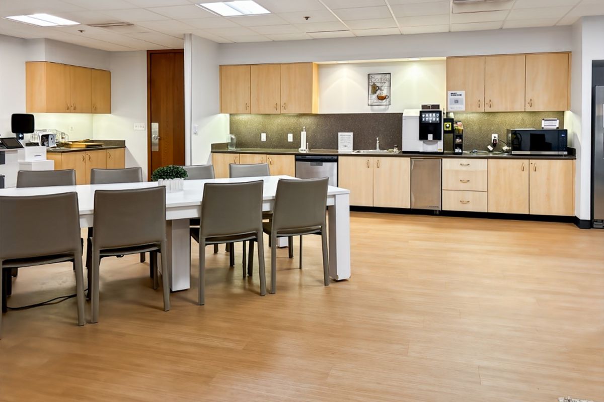 A spacious open-plan office breakroom featuring light wood cabinetry, integrated sink, modern laminate flooring, and a functional kitchenette area with recessed lighting.