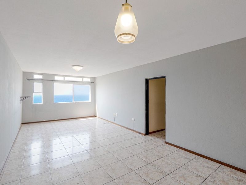 An empty, light-filled living area featuring tiled flooring, neutral walls, and a window with a view of the ocean.
