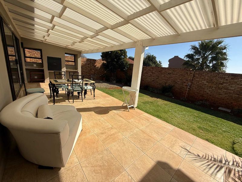Covered patio area featuring tiled flooring, a built-in braai, and a pergola roof, overlooking a private garden with a brick boundary wall.