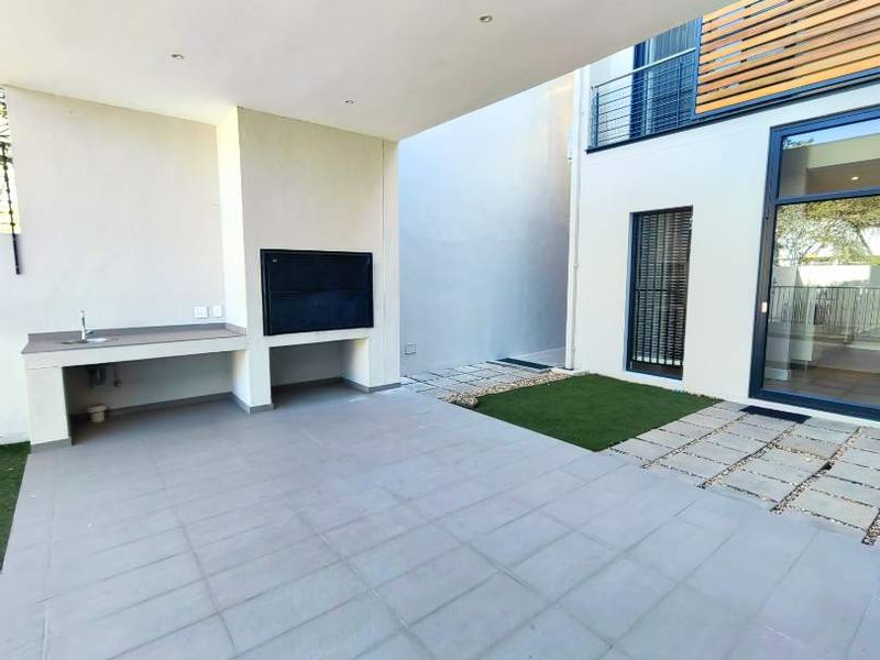 A modern covered patio featuring a built-in braai, outdoor sink, tiled flooring, and direct access to a small garden area with stone pavers.