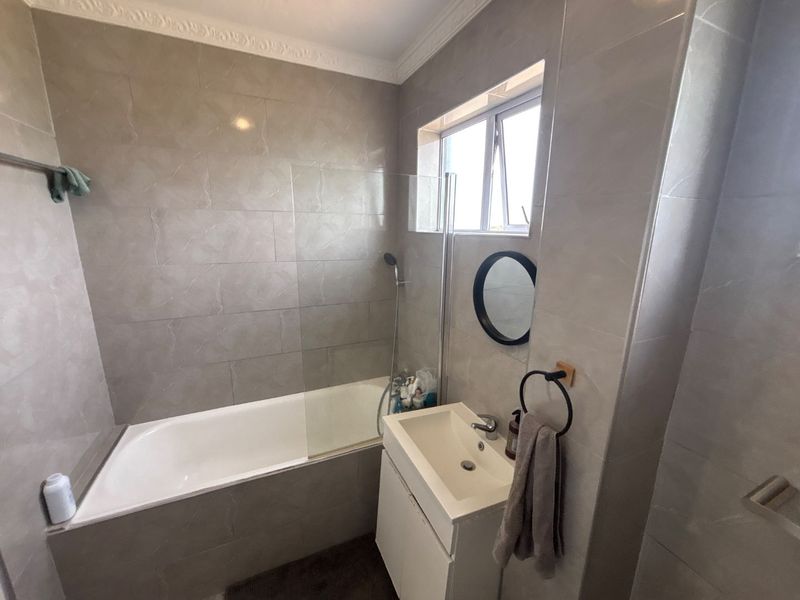 A modern bathroom featuring full-height wall tiling, a built-in bathtub with a glass shower screen, and a compact wall-mounted vanity unit.