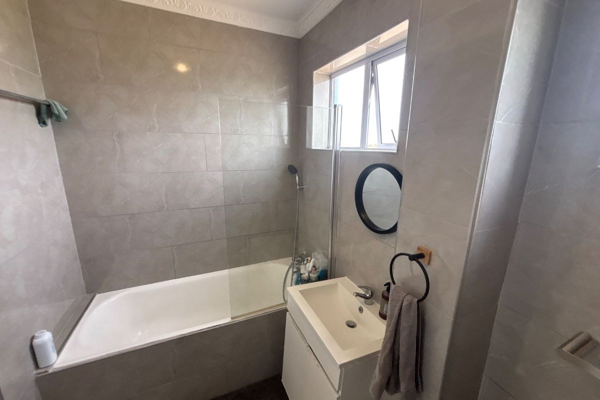 A modern bathroom featuring full-height wall tiling, a built-in bathtub with a glass shower screen, and a compact wall-mounted vanity unit.