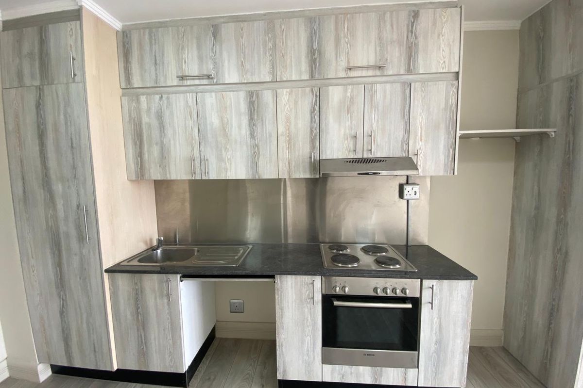 A compact kitchen featuring wood-look cabinetry, a stainless steel splashback, integrated electric hob, oven, and extractor fan with a single basin sink.