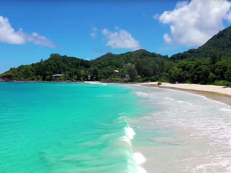 A scenic aerial view of a pristine tropical beach with turquoise waters, white sand, and lush green hills in the background.
