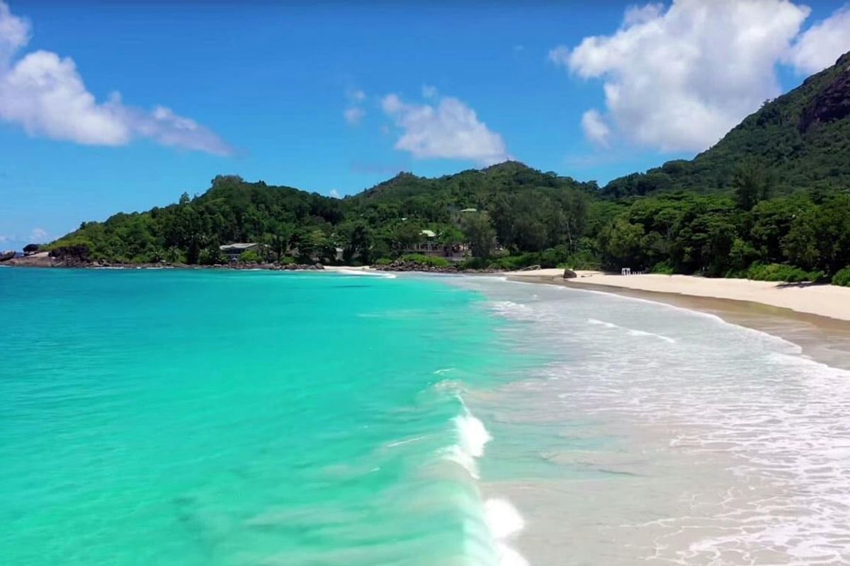 A scenic aerial view of a pristine tropical beach with turquoise waters, white sand, and lush green hills in the background.