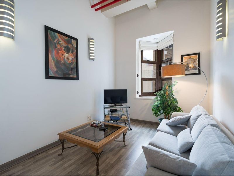 A well-lit living room featuring laminate flooring, modern wall-mounted lighting, and an exposed industrial-style ceiling pipe detail.