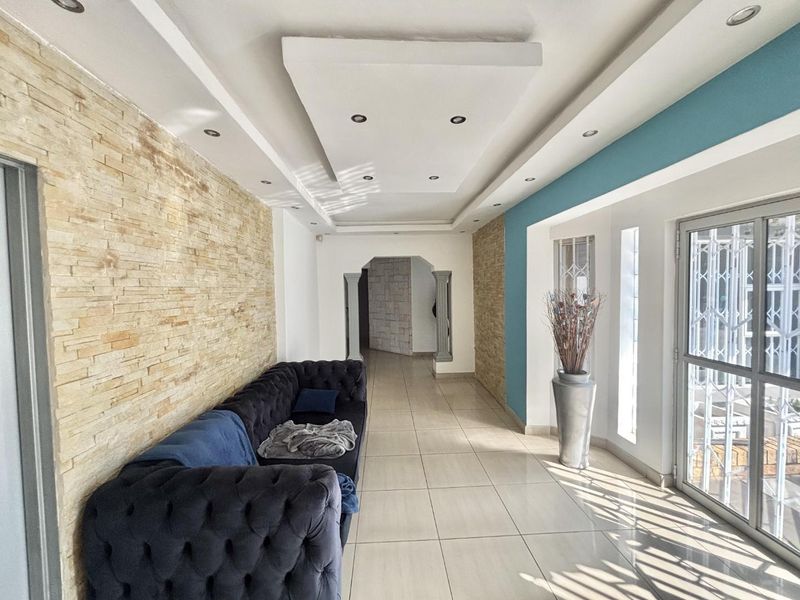 Spacious living area featuring a textured stone accent wall, recessed ceiling lighting, and large windows with security gates, finished with neutral floor tiles.