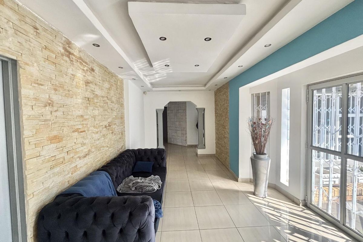 Spacious living area featuring a textured stone accent wall, recessed ceiling lighting, and large windows with security gates, finished with neutral floor tiles.