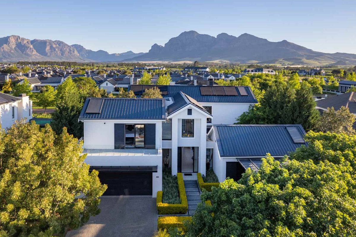 Aerial view of a modern double-story residence featuring a metal roof with solar panels, a paved driveway, and a landscaped garden.