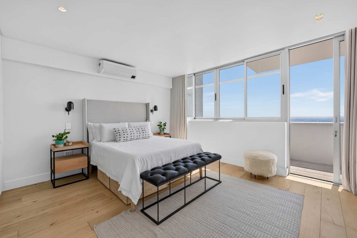 A bright and modern bedroom featuring timber flooring, a split-system air conditioner, and direct access to a private balcony with ocean views.