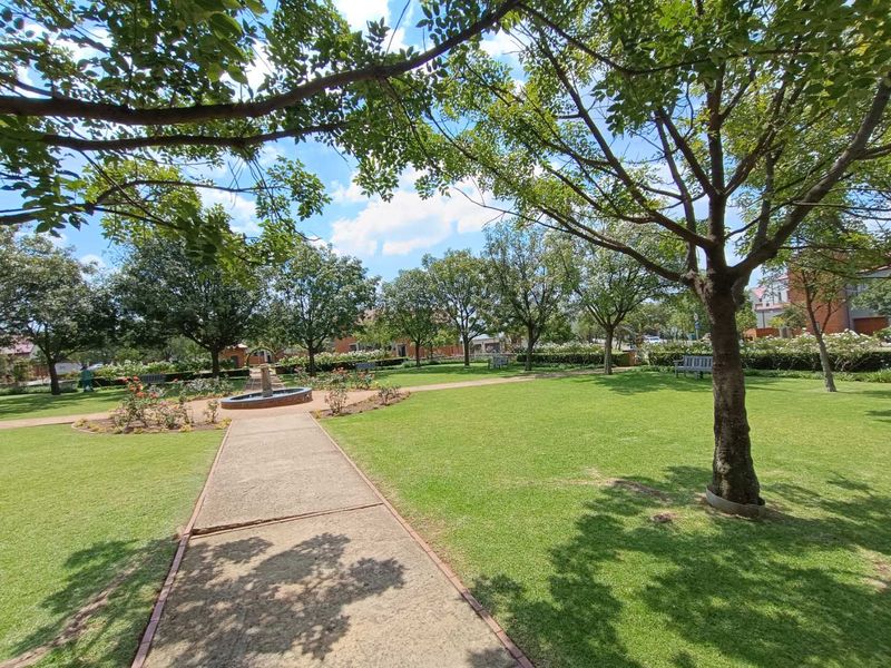 A well-maintained communal garden area featuring lush green lawns, mature trees, a central water fountain, and paved walkways.