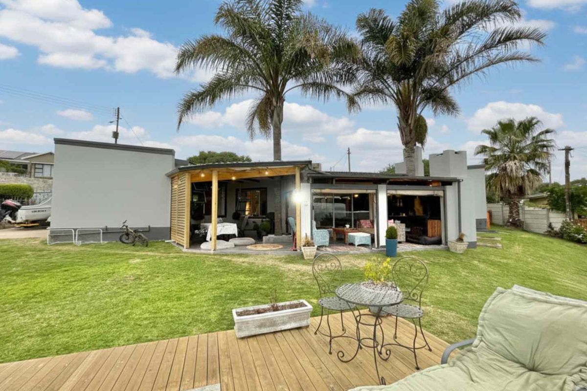 Rear view of the property featuring a covered patio area, timber decking, and a well-maintained garden with mature palm trees.