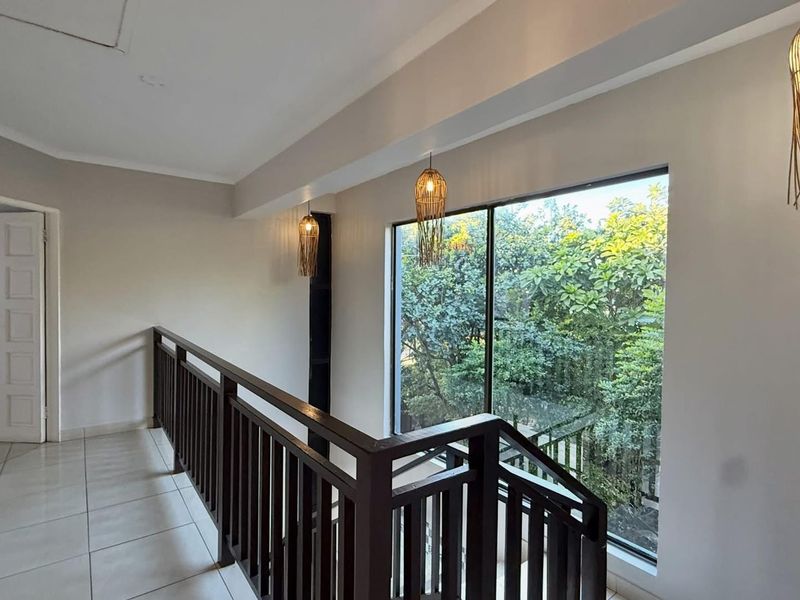 A bright and spacious upper-level landing featuring a wooden balustrade, tiled flooring, and a large window overlooking lush greenery.