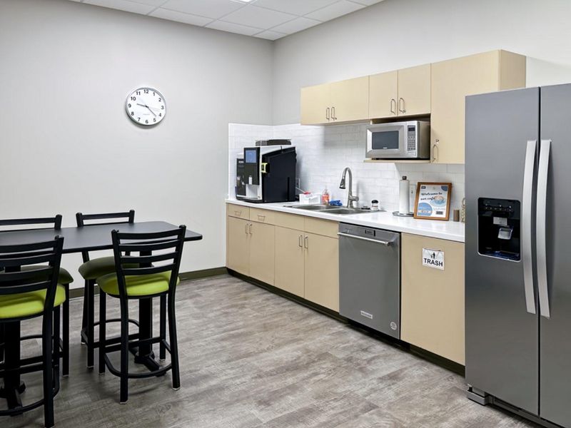 A clean office kitchenette featuring light wood cabinetry, a stainless steel sink, integrated dishwasher, built-in microwave, and a subway tile backsplash.