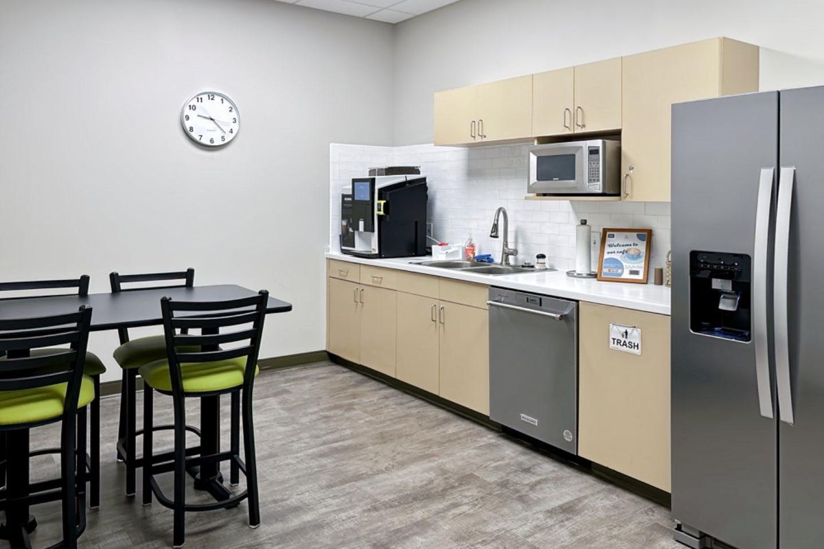 A clean office kitchenette featuring light wood cabinetry, a stainless steel sink, integrated dishwasher, built-in microwave, and a subway tile backsplash.