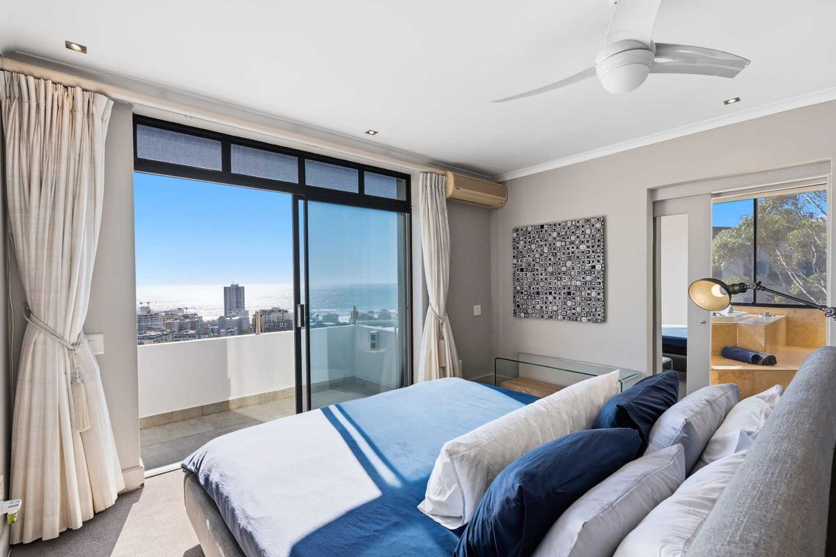 A bright bedroom featuring a large sliding glass door leading to a balcony with expansive ocean and city views, complemented by a ceiling fan.