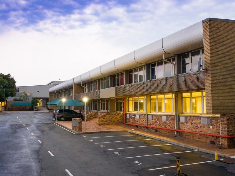 Exterior view of a multi-story industrial office building featuring a brick facade, aluminum window frames, and an adjacent paved parking area with covered bays.