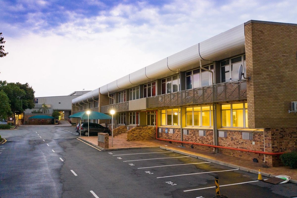 Exterior view of a multi-story industrial office building featuring a brick facade, aluminum window frames, and an adjacent paved parking area with covered bays.