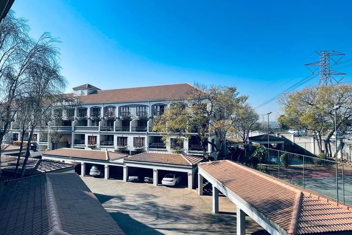An elevated view of the apartment complex featuring multiple levels of balconies, covered parking structures, and paved common areas under a clear sky.
