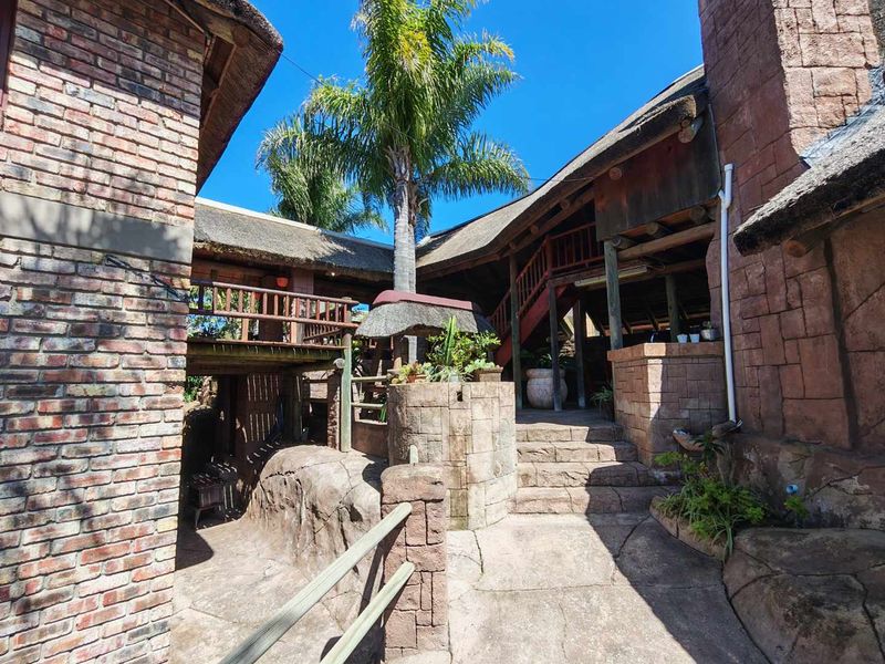 A rustic rear exterior view featuring thatched roofing, brick and stone masonry, a timber staircase, and a raised balcony area.