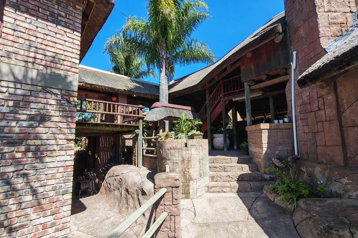 A rustic rear exterior view featuring thatched roofing, brick and stone masonry, a timber staircase, and a raised balcony area.