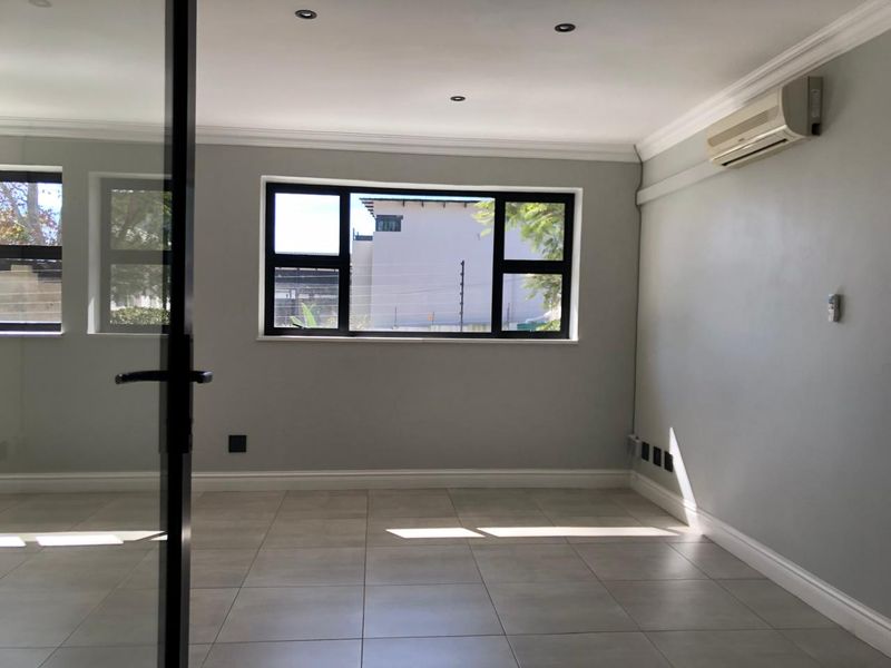 A bright, empty office space featuring tiled flooring, recessed downlights, a wall-mounted air conditioning unit, and modern black-framed windows.